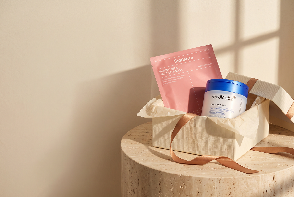 Open ivory gift box with a Biodance collagen mask and a medicube Zero Pore Pad jar on a travertine plinth